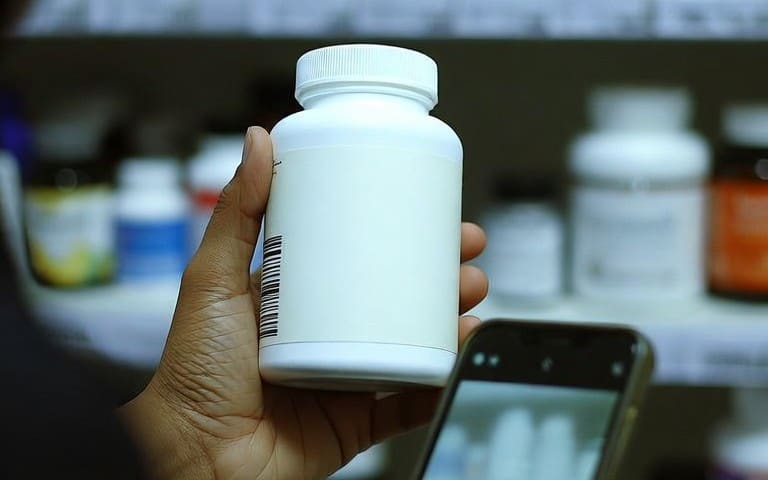 يد تمسك علبة مكمّل بيضاء بلا اسم يظهر على جانبها الباركود، وأمامها جوال بكاميرا مفتوحة، أمام رفوف مكمّلات في صيدلية
